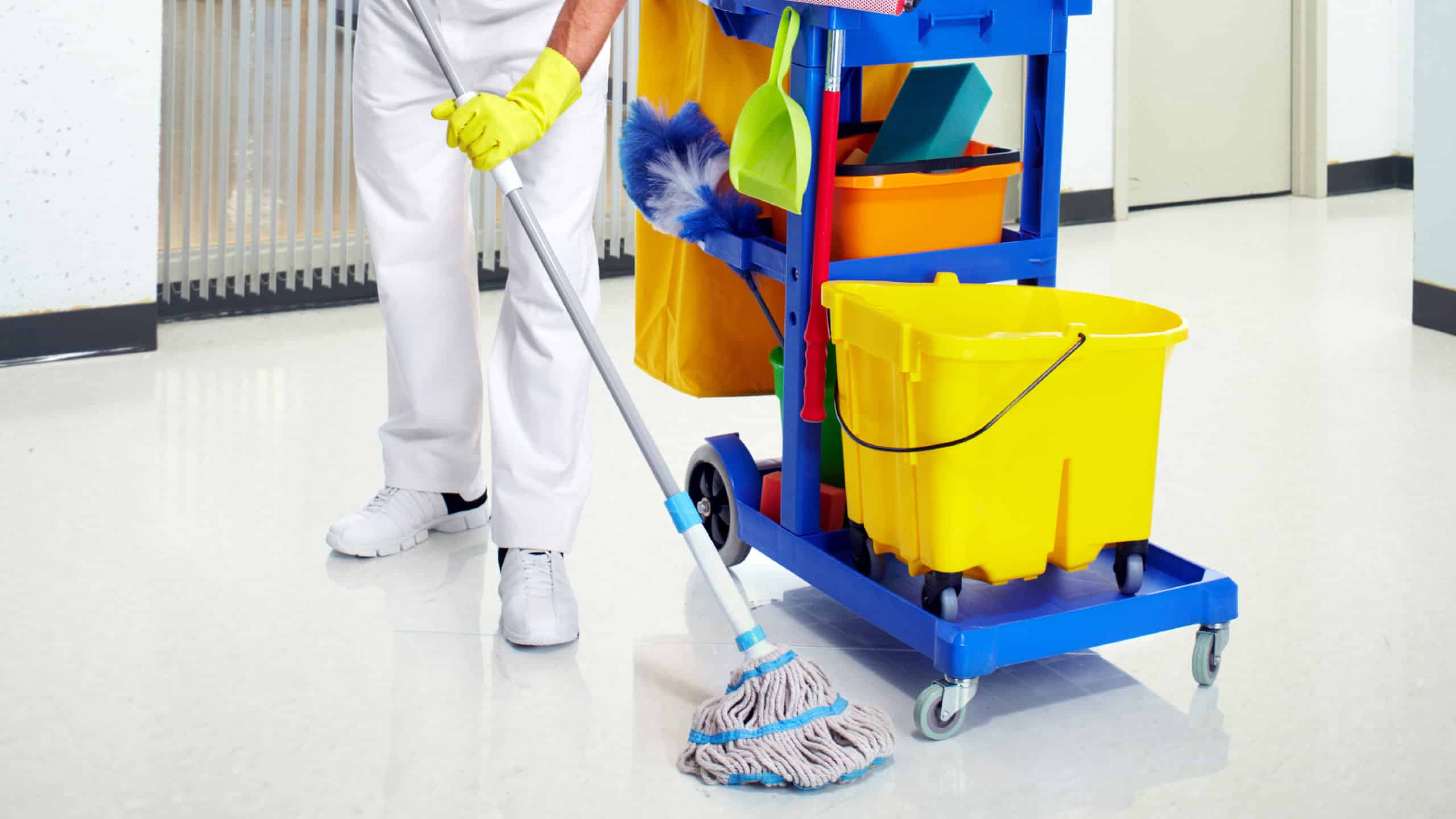A school janitor wipes a linoleum floor with a traditional mop, a janitorial cart with a bucket of water stands nearby.